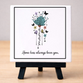 Small framed artwork with floral design and text on a white ceramic tile resting on a small black easel